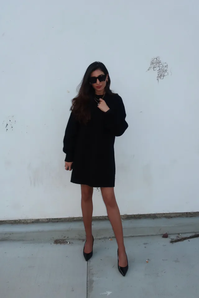 Ayman in a black dress and sunglasses against a pale wall.
